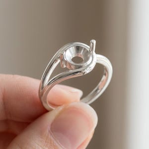 May include: A polished silver ring setting featuring a circular central mount with two vertical prongs designed to hold a gemstone. The band has a smooth, reflective finish and a split shank design that curves upward to support the setting.