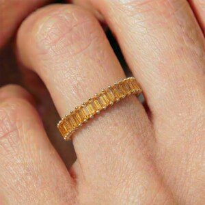 May include: A gold band ring featuring a continuous row of rectangular, baguette-cut yellow gemstones set in a channel. The stones are arranged side-by-side, creating a uniform, linear pattern that encircles the finger with a bright, warm hue.