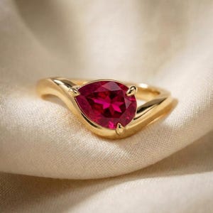 Op de afbeelding: Een gouden ring met een enkele peervormige, dieprode edelsteen gezet in een gepolijste geelgouden band. De steen wordt vastgehouden door drie pootjes en de band heeft een glad, gebogen ontwerp dat taps toeloopt naar de zetting van de rode edelsteen.