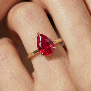 May include: A gold ring featuring a single pear-shaped, deep red gemstone set in a four-prong mounting. The band is smooth and polished, showcasing a classic design that highlights the vibrant color and faceted cut of the central stone.