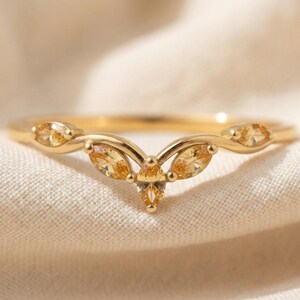 May include: A gold V-shaped ring featuring five marquise-cut yellow gemstones arranged in a curved pattern along the band. The metal has a polished finish, and the stones are set securely within the gold frame, creating a symmetrical design.