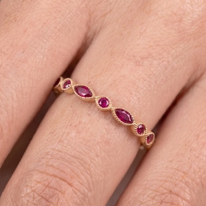 Puede incluir: Un anillo de oro con un patrón alterno de rubíes de color rojo intenso en talla marquesa y redonda. El engaste de metal presenta una delicada textura de cuentas a lo largo de los bordes, creando un borde decorativo continuo alrededor de cada gema.
