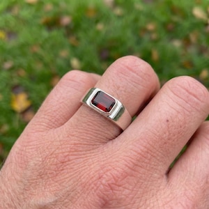 Puede incluir: Un anillo de plata con una piedra preciosa rectangular de color rojo intenso. El anillo tiene un diseño sencillo y moderno con un acabado pulido. La piedra preciosa está engastada en un bisel.
