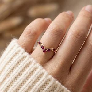 Könnte beinhalten: Ein goldener Ring mit geschwungenem Design, besetzt mit mehreren kleinen, oval geformten roten Edelsteinen. Der Ring wird an einem Finger getragen, der Hintergrund ist weich verschwommen. Das Design des Rings ist zart und elegant.