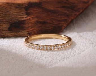 May include: A thin gold band ring featuring a continuous row of small, clear, round-cut stones set along the outer circumference. The metal has a polished yellow finish, and the stones are arranged in a pavé style, creating a uniform, sparkling look.
