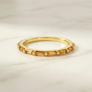 May include: A gold band ring featuring a series of alternating rectangular baguette-cut amber-colored gemstones and small polished gold spheres set along the entire circumference of the thin metal band.