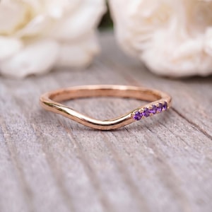 Op de afbeelding: Een roségouden ring met een gebogen band, voorzien van een rij van vijf kleine, ronde, paarse amethist edelstenen aan één kant van de boog. Het metaal heeft een gepolijste, reflecterende afwerking en de band volgt een golvende vorm.