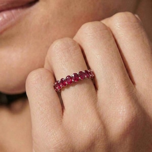 May include: A silver ring featuring a row of oval-cut, vibrant red gemstones. The ring is worn on a finger, showcasing the gemstones' rich color and sparkle. The ring is a piece of jewelry.