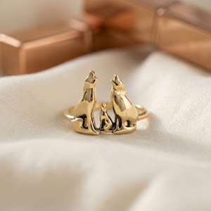 May include: A gold-toned ring featuring a sculptural design of two adult wolves howling and a smaller wolf pup positioned between them. The metal has a polished finish, and the figures are integrated into the band of the ring.