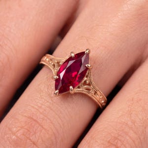 May include: A rose gold ring featuring a marquise-cut deep red gemstone as the centrepiece. The band displays intricate, ornate metalwork with swirling patterns along the sides, held securely by six prongs surrounding the central stone.
