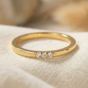 May include: A polished gold band ring featuring three small, round, clear stones set horizontally in a row along the top of the band. The metal has a smooth, reflective finish, and the stones are evenly spaced and embedded within the gold surface.