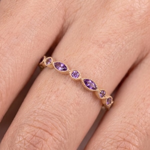 Op de afbeelding: Een ring van roségoud met een afwisselend patroon van marquise- en rondgeslepen paarse amethisten. De metalen zetting is voorzien van een verfijnde milgrain-textuur langs de randen, wat zorgt voor een doorlopend decoratief ontwerp.