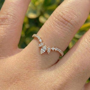 May include: A rose gold ring featuring a V-shaped band adorned with a central marquise-cut diamond, flanked by smaller round-cut diamonds and two additional marquise-cut diamonds, creating a delicate, symmetrical, and pointed chevron design.