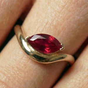 Puede incluir: Un anillo de oro con una piedra preciosa de color rojo intenso en talla marquesa engastada horizontalmente. La banda tiene un acabado liso y pulido con un diseño curvo que sigue la forma de la piedra, creando un aspecto elegante y continuo.