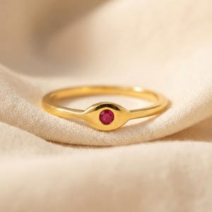 May include: A gold ring featuring a smooth, polished band and a central eye-shaped setting. The center of the eye motif contains a small, round, vibrant red gemstone. The ring has a consistent metallic finish throughout the entire band and setting.