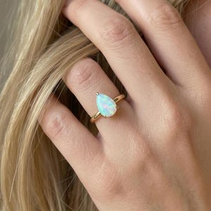 May include: A gold ring featuring a pear-shaped opal gemstone set in a simple prong setting. The opal displays iridescent flashes of color, including blue, green, and pink, against a milky white base. The band is a smooth, polished yellow gold.