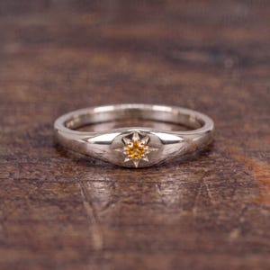 May include: A polished silver-toned ring featuring a central starburst setting that holds a small, round, vibrant yellow gemstone. The band is smooth and tapers slightly toward the front, where the star-shaped engraving surrounds the stone.