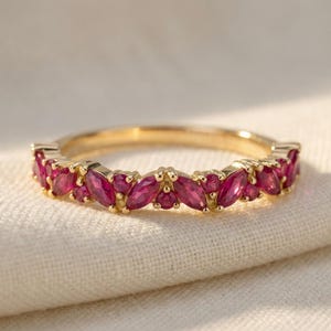 May include: A gold band ring featuring a series of alternating marquise-cut and round-cut vibrant pink gemstones set along the top half of the band. The metal has a polished finish, and the stones are arranged in a repeating pattern across the front.