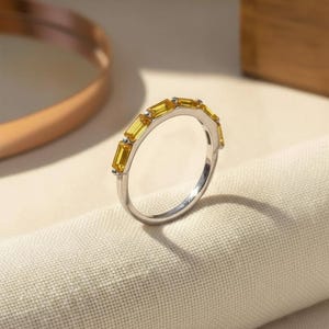 Puede incluir: Un anillo de metal plateado que presenta una serie de cinco piedras preciosas amarillas rectangulares, con talla baguette, dispuestas a lo largo de la mitad superior de la banda. Las piedras están alineadas horizontalmente con pequeñas garras.