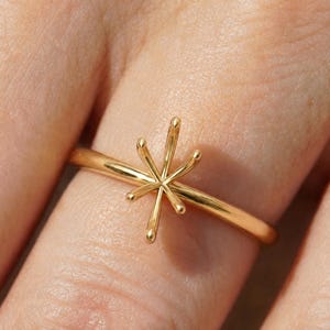 May include: A polished gold ring featuring a six-prong starburst setting designed to hold a gemstone. The band is thin and smooth, with the prongs extending outward in a symmetrical star shape, creating a clean and structural metal ring base.