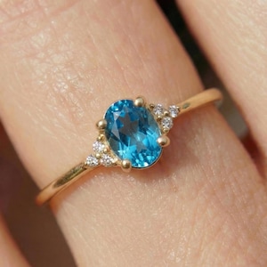 May include: A gold ring featuring a central oval-cut blue gemstone held by four prongs. The band is thin and polished gold. On each side of the central stone, there are three small, clear, round-cut accent stones set into the gold metal band.