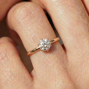 May include: A solitaire ring featuring a round brilliant-cut clear gemstone set in a polished rose gold band. The center stone is held by six prongs, showcasing a high-clarity faceted surface that reflects light with a bright, sparkling appearance.