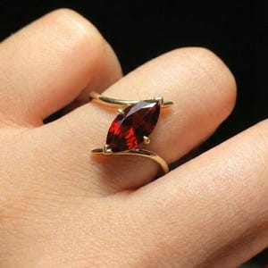 May include: A gold ring featuring a marquise-cut, deep red gemstone. The gemstone is set in a gold band that curves around the finger. The ring is displayed against a dark background, highlighting the vibrant color of the stone.
