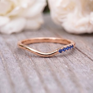 Könnte beinhalten: Ein geschwungener Ring aus Roségold mit einer Reihe von fünf kleinen, runden, tiefblauen Saphiren, die an einer Seite des Bandes eingefasst sind. Das Metall hat eine polierte Oberfläche und der Ring weist eine sanfte Wellenform auf.