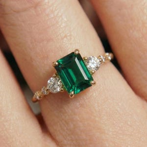 May include: A gold ring featuring a central emerald-cut green gemstone flanked by two round brilliant-cut white diamonds on each side. The band is thin and polished, showcasing a classic prong setting that secures the stones in place.