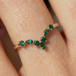 Pode incluir: Um anel de ouro rosa com um design curvo, adornado com múltiplas pedras preciosas verde esmeralda. As pedras preciosas têm várias formas, incluindo cortes redondos e marquesa, criando uma aparência delicada e elegante.