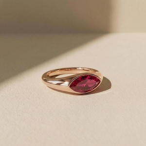 May include: A rose gold ring featuring a single marquise-cut deep pink gemstone set horizontally in a smooth, polished metal band. The ring has a sleek, solid construction with a tapered design that highlights the vibrant color of the stone.