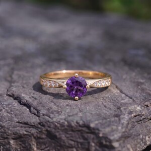 Op de afbeelding: Een gouden ring met een ronde, gefacetteerde paarse amethist in het midden, vastgehouden door vier pootjes. De ringband is gemaakt van gepolijst goud en is aan beide zijden bezet met kleine, heldere, rondgeslepen witte steentjes.