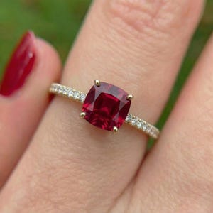 May include: A gold ring featuring a large, cushion-cut, deep red gemstone set in four prongs. The slender band is accented with a row of small, clear, round-cut stones embedded along the shank on both sides of the central red gemstone.