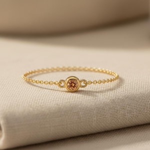 Puede incluir: Un delicado anillo de cadena dorada con una piedra preciosa redonda de color melocotón claro engastada en un bisel dorado liso. El anillo está formado por una fina cadena de eslabones que conecta con el engaste central por ambos lados.