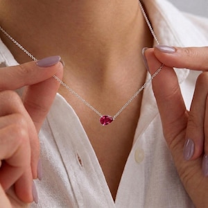 May include: A delicate silver chain necklace featuring a single, faceted, pear-shaped ruby-colored gemstone pendant. The pendant hangs centrally from the thin metallic chain, which rests against a white collared shirt.