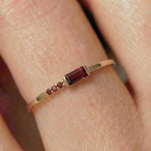 May include: A delicate gold ring featuring a rectangular, deep red gemstone as the focal point, flanked by three smaller, round red stones. The ring's band is slender and polished, creating a minimalist and elegant design.