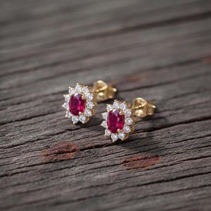 May include: A pair of gold stud earrings, each featuring a central oval-cut red ruby surrounded by a halo of small, round, clear white diamonds. The earrings have a post-back closure and are crafted from polished yellow gold metal.