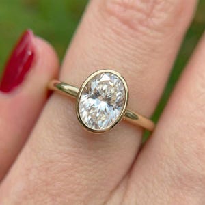 May include: A gold ring featuring an oval-cut diamond. The diamond is set in a bezel setting, and the band is a thin, polished gold. The ring is shown on a finger, with a blurred green background.