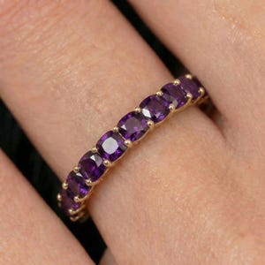 May include: A gold band ring featuring a continuous row of cushion-cut purple amethyst gemstones. Each gemstone is held in place by small gold prongs, creating a uniform and repeating pattern along the entire circumference of the metal ring band.