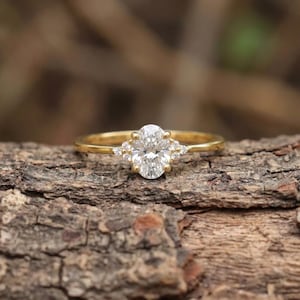 May include: A gold ring featuring a central oval-cut clear gemstone held by four prongs, flanked by three small round-cut clear stones on each side. The band is smooth and polished, showcasing a classic design with a bright, reflective finish.