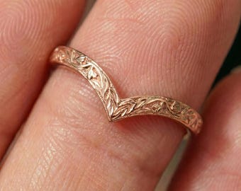 May include: A rose gold V-shaped ring featuring an intricate engraved leaf pattern along the entire outer band. The metal has a polished finish that catches the light, highlighting the detailed botanical carvings etched into the surface of the band.