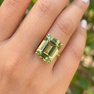 May include: A gold ring featuring a large, rectangular-cut, light green gemstone. The gemstone is set with gold prongs. The ring is worn on a finger.