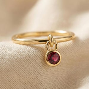May include: A gold ring featuring a thin, smooth band and a single, small, circular charm dangling from a jump ring. The charm is set with a round, deep red gemstone, securely held in a polished gold bezel setting that matches the band's finish.