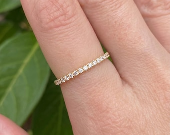 May include: A thin gold band ring featuring a single row of small, round, clear faceted stones set along the top half of the band. The metal has a warm yellow tone, and the stones are arranged in a continuous line, creating a delicate, sparkling appearance.
