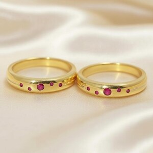 May include: Two gold rings featuring a polished finish and a series of small, round, flush-set red gemstones arranged in a cluster along the outer band. Each ring displays one larger central red stone flanked by several smaller red accent stones.