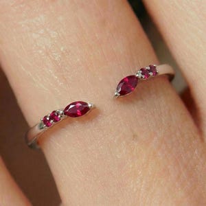 May include: An open-style silver-toned ring featuring two marquise-cut red rubies and four smaller round-cut red rubies. The stones are set along the two ends of the open band, creating a symmetrical design with vibrant red accents on a metallic base.