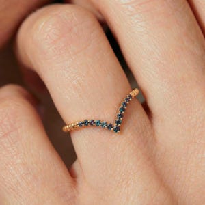 May include: A V-shaped gold ring featuring a row of small, round, multi-colored gemstones that shift between shades of blue, purple, and teal. The band has a polished gold finish and follows a chevron silhouette along the finger.