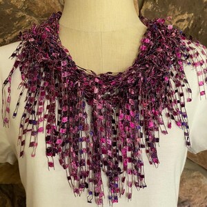 May include: A purple and pink crocheted fringe necklace with a square pattern. The necklace is on a white mannequin.
