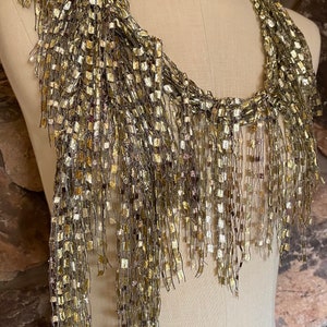 May include: A gold and brown fringed necklace made of woven metallic thread. The necklace is displayed on a white mannequin.
