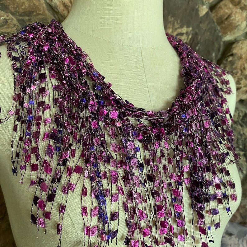 May include: A handmade necklace with a fringe design. The necklace features a variety of square-shaped beads in shades of purple, pink, and blue. The beads are attached to a black thread, creating a textured and layered effect. The necklace is displayed on a mannequin.